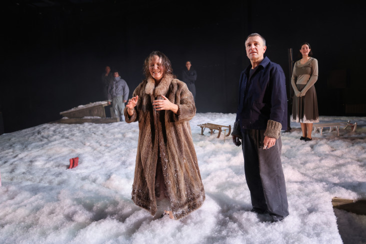 Eine Schauspielerin in einem Pelzmantel steht neben einem Schauspieler. Der Bühnenboden ist mit Schnee bedeckt. Im Hintergrund stehen Schlitten.