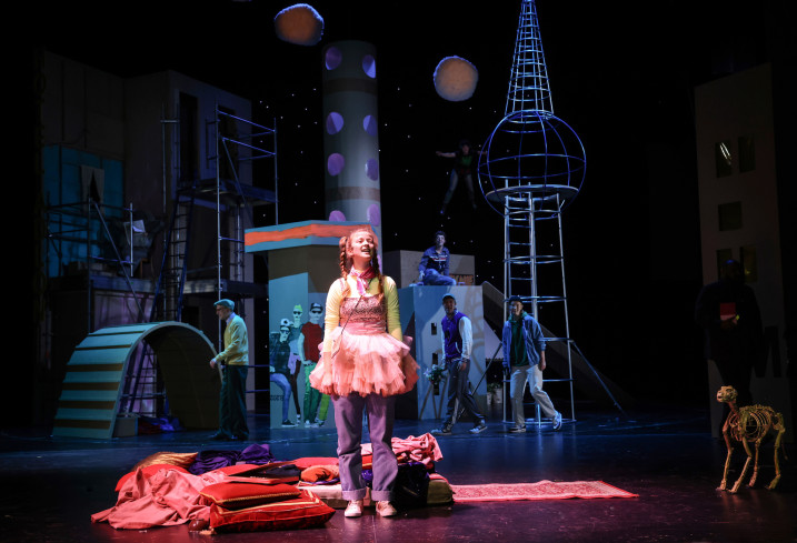 Vor dem dunklen Hintergrund der Stadt steht eine Person vorn an der Bühne im Licht. Sie trägt ein rosafarbenes Kleid. 