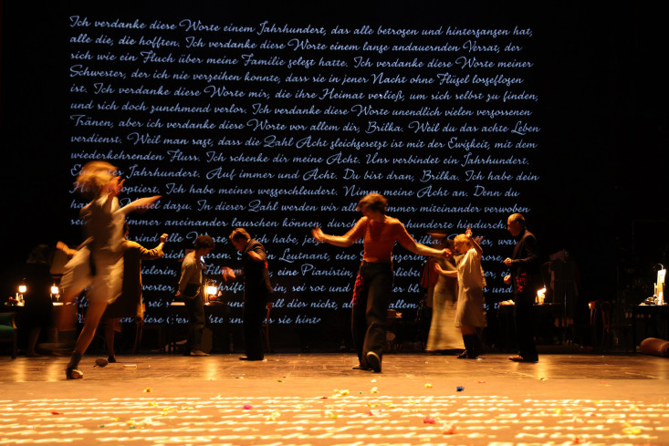 Auf der Bühnenrückseite ist ein langer Text in weißer Schrift zu sehen. Auf der Bühne tanzt das Ensemble. Auch auf dem Bühnenboden ist weiße Schrift.