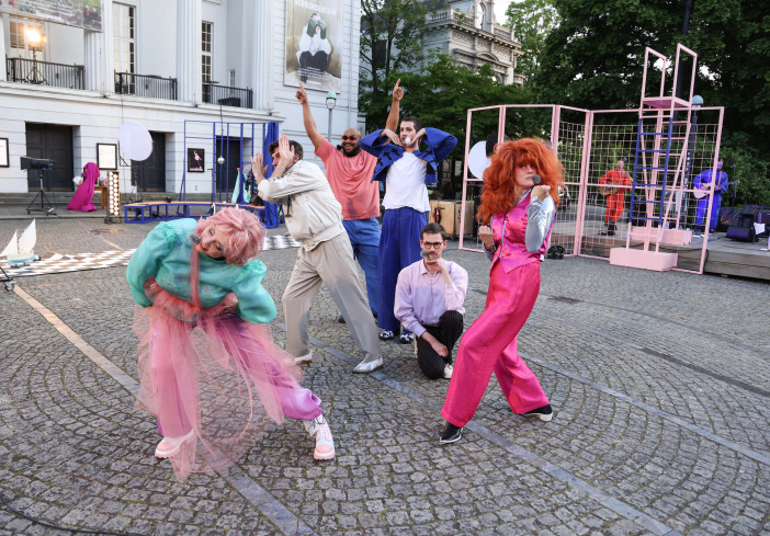 Sechs Personen stehen auf dem Goetheplatz. Fast alle sind sehr auffällig und farbenfroh gekleidet. Sie tanzen und singen. Im Hintergrund spielt eine Band.