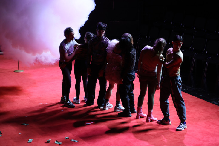 Sieben Personen stehen in einer Reihe auf dem roten Teppich. Sie lehnen aneinander. Hinten auf der Bühne ist Nebel.