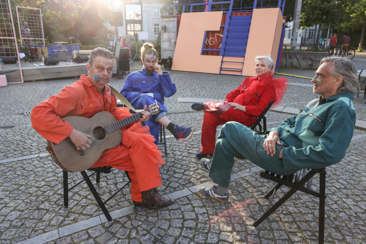 Vier Menschen sitzen auf Klappstühlen auf dem Goetheplatz. Sie tragen alle bunte Overalls. Einer hält eine Gitarre in der Hand.