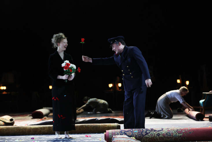 Ein Schauspieler in einer militärischen Uniform übergibt eine rote Rose an eine lachende Person, die schon einige Blumen in der Hand hält. Im Hintergrund rollen andere Teppiche zusammen.
