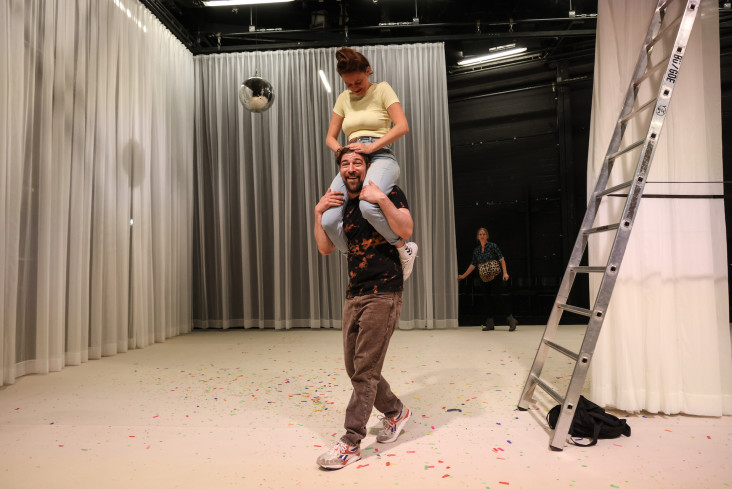 Ein Schauspieler trägt eine Schauspielerin auf seinen Schultern. Neben ihnen steht eine sehr hohe Leiter. Auf dem Teppich liegt lauter Konfetti. 