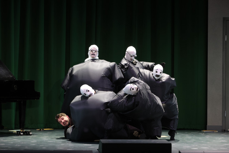 Fünf Personen in dunklen, aufgeblasenen Kostümen stehen eng beieinander. Sie halten einen Sänger auf den Boden gedrückt. Nur sein Kopf ist zu sehen.
