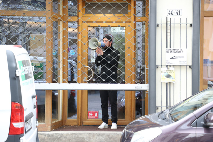 Ein Mann steht mit einem Megafon im Eingang zu einem Geschäft. Vor ihm öffnet sich das Gitter, das die Schaufensterscheiben schützt, wenn der Laden geschlossen ist. Autos parken vor dem Laden.