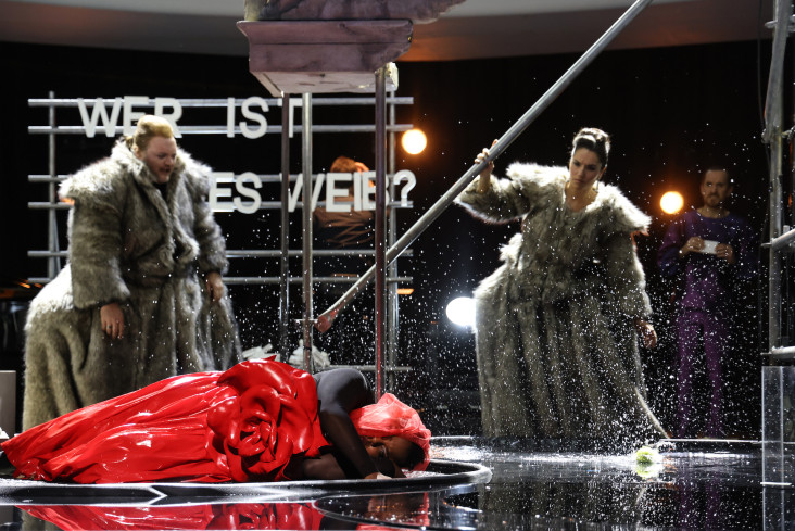 Eine Sängerin in einem roten Kleid liegt in einem Wasserbecken. Hinter ihr stehen ein Sänger und eine Sängerin. Beide tragen ausladende Pelzmäntel. 