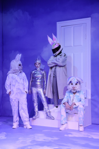 Vor einer weißen Tür ist eine weiße Treppe. Auf der stehen und sitzen vier Personen in aufwändigen Kostümen. Drei sind Hasen, eine andere trägt einen silbernen Anzug.