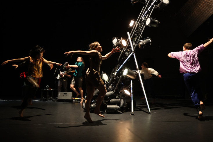 In der Mitte der Bühne steht eine Installation aus Scheinwerfern. Um sie herum tanzen fünf Personen. Alle schwingen die Arme in der Luft. 