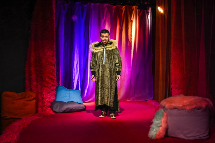 Ein junger Akteur steht in einem bodenlangen goldenen Mantel in der plüschigen und knallbunten Bühnenumgebung.