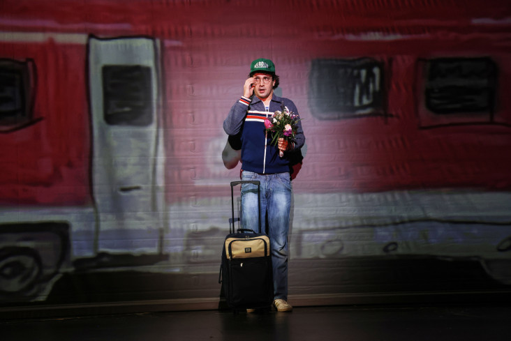 Ein Schauspieler mit einem Rollkoffer steht auf der Bühne. Hinter ihm ist ein gemalter Zug.