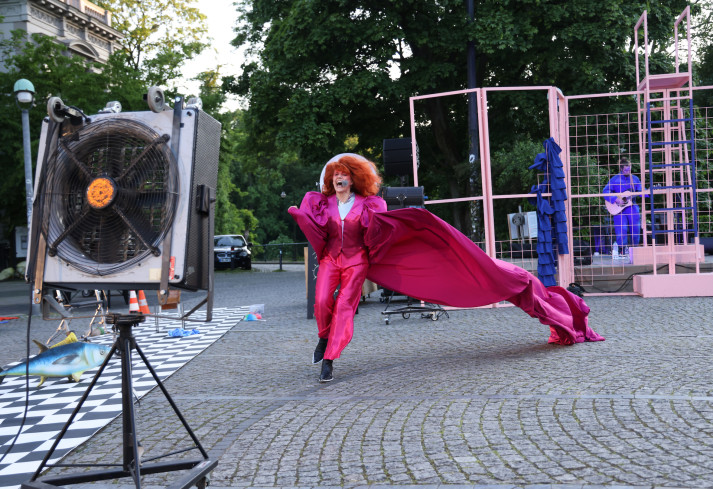 Eine Schauspielerin läuft auf eine Windmaschine zu. Sie trägt eine große, rote Perücke. Ihr langer, pinker Mantel fliegt im Wind.
