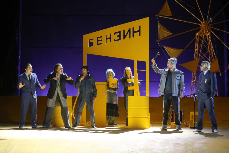Sieben Personen stehen auf der Bühne. Zwischen ihnen steht eine gelbe Tankstellen-Kulisse. Im Hintergrund ist ein großes Windrad.
