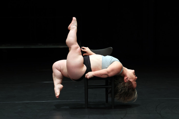 Eine Tänzerin liegt mit dem Rücken auf einem Stuhl und streckt ein Bein senkrecht nach oben, das andere ist angewinkelt. 