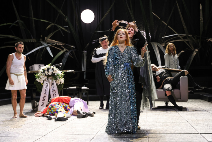 Vorn an der Bühne steht eine Sängerin in einem langen, blauen, glitzernden Kleid. Die Person hinter ihr setzt ihr eine Krone auf. Drei andere Personen schauen zu. Auf dem Boden liegen zwei weitere Menschen. 