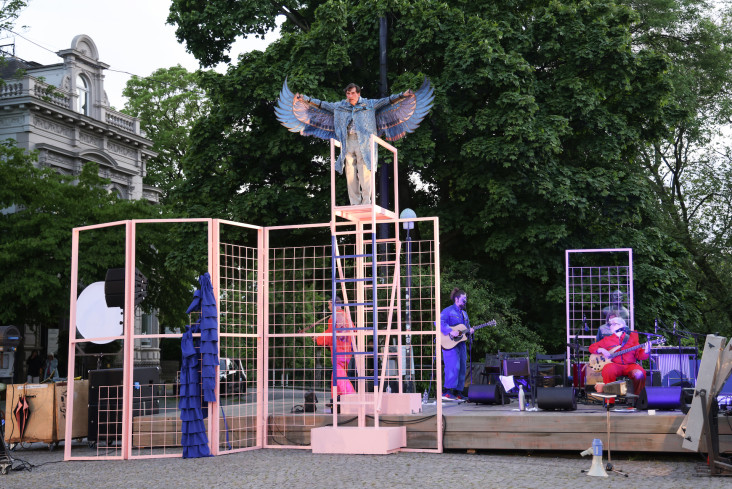 Auf einer Bühne auf dem Goetheplatz steht oben auf einer Leiter ein Schauspieler. Er trägt ein Kostüm mit großen, blauen Flügeln. Unten stehen andere, eine Band macht Musik.