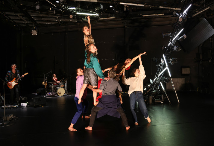 In der Mitte der Bühne bilden sechs Tänzerinnen und Tänzer eine menschliche Skulptur. Dabei werden einige von den anderen in der Luft gehalten. Hinten steht die Band.
