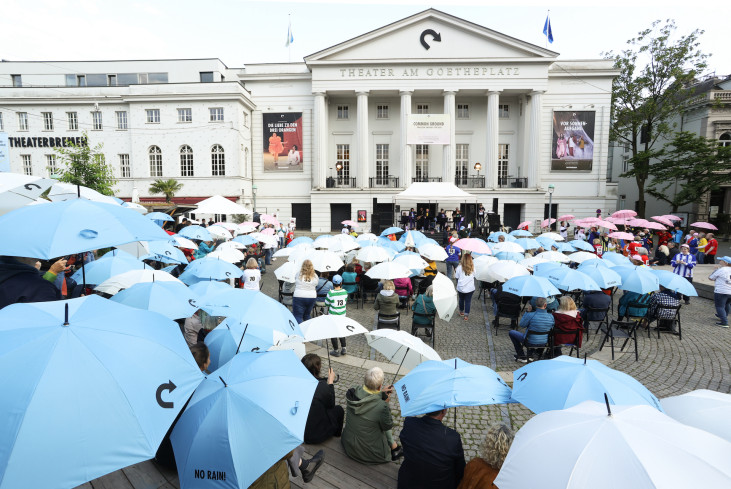 Sehr viele Menschen sitzen mit aufgespannten Regenschirmen auf dem Goetheplatz. Es regnet nicht. Im Hintergrund ist das Theater.