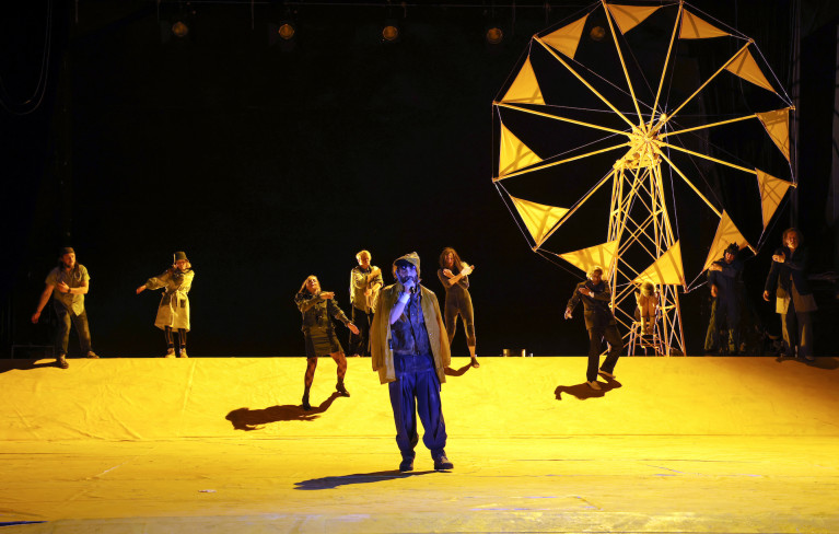 Der Bühnenboden leuchtet gelb. Im Hintergrund steht ein gelbes Windrad. Vorn steht ein Schauspieler. Hinter ihm verteilt sind neun Personen. Manche tanzen.