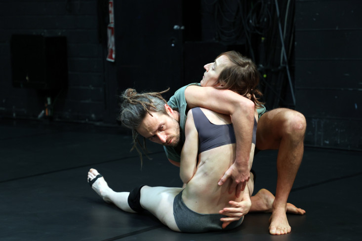 Ein Tänzer hält eine auf dem Boden sitzende Tänzerin im Arm. Er hat seine Hände auf ihrem Rücken. 