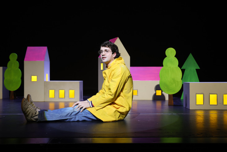 Ein Schauspieler in einer gelben Regenjacke sitzt auf der Bühne. Hinter ihm stehen kleine Häuser. Die Fenster sind erleuchtet.