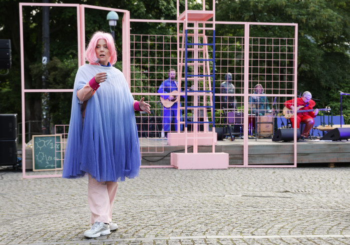 Eine Schauspielerin mit einer rosanen Perücke steht auf dem Goetheplatz. Sie trägt ein blaues Kleid. Hinter ihr ist das Bühnenbild aus rosa Gittern. Eine Band macht Musik.