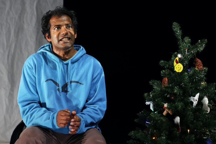 Der Schauspieler sitzt neben einem kleinen Tannenbaum und spricht. Er trägt einen hellblauen Kapuzenpullover auf dem mit schwarzer Farbe ein Vogel aufgezeichnet ist.