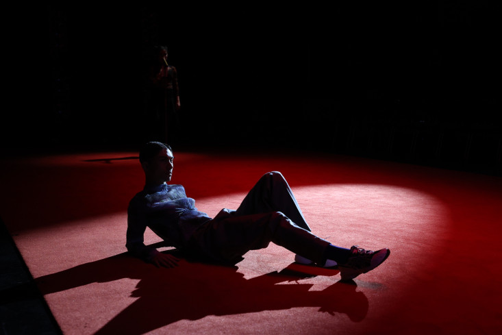 In einem Lichtkegel auf der Bühne liegt ein Tänzer auf dem roten Teppich.