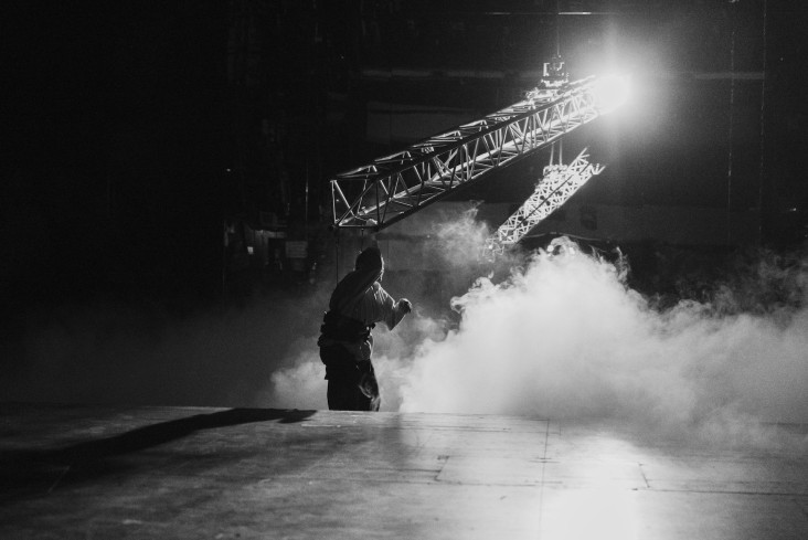 Eine Person hängt an einem großen Pendel über der Bühne. Sie schwebt. Auf dem Boden ist Nebel. Nur ein Scheinwerfer leuchtet.