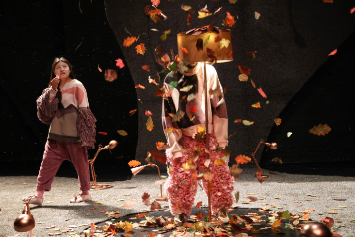 Ein Schauspieler steht in der Mitte der Bühne, sein Gesicht wird von einer Stehlampe verdeckt. Blätter fallen auf ihn herunter. Hinten geht eine Schauspielerin. Sie schaut ihn an.