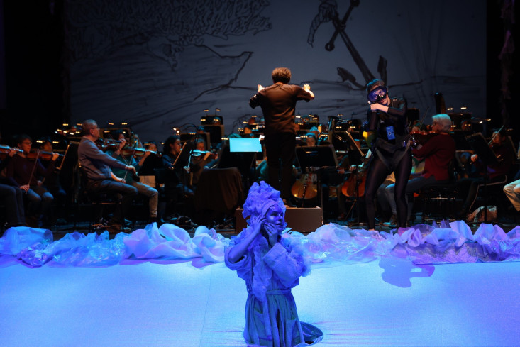 Die Bühne ist blau beleuchtet. Am Rand vorn taucht eine Sängerin aus dem Bühnenboden auf. Sie trägt einen blauen Bademantel und eine blaue Kopfbedeckung. Hinten ist das Orchester.