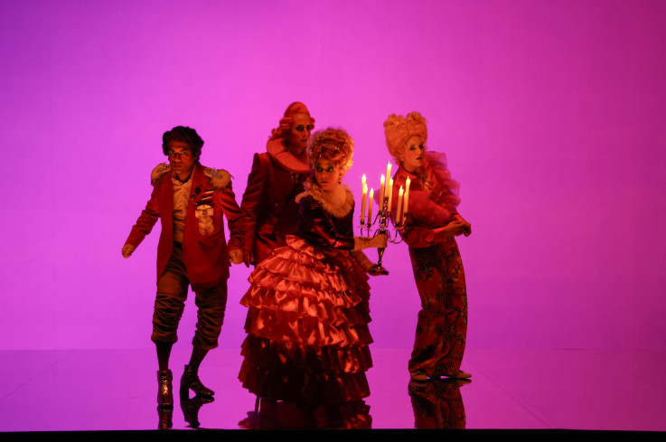 Vier Personen stehen eng beieinander. Alle tragen Kostüme, die ans 18. Jahrhundert erinnern, aber sehr bunt sind. Eine Schauspielerin in einem Reifrock hält einen Kerzenleuchter. Der Hintergrund und das Licht sind pink.