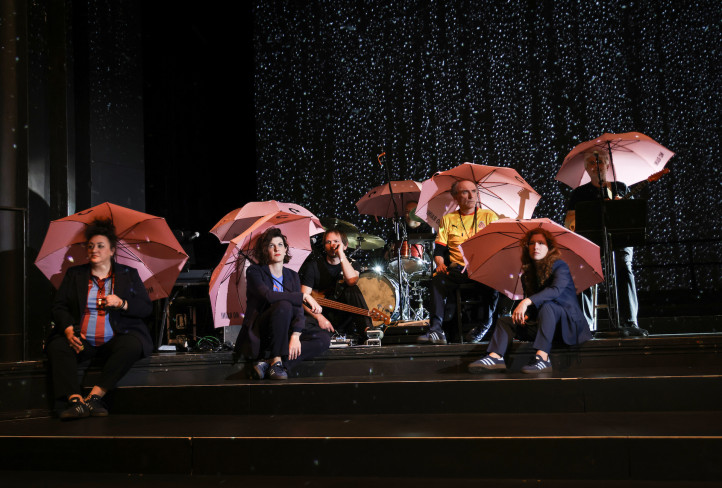 Auf der Bühne ist es dunkel. Sieben Personen sitzen auf Stufen. Alle haben rosafarbene Regenschirme aufgespannt. 