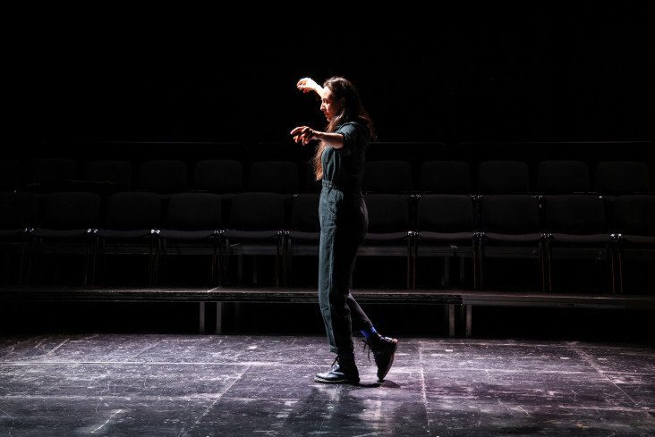 Eine Frau tanzt allein auf der großen Bühne. Ihre Arme sind zur Seite erhoben. Sie trägt einen schwarzen Overall und hat langes, dunkles Haar. 