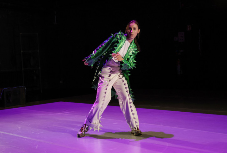 Eine Schauspielerin tanzt. Sie trägt eine weiße Hose und eine grüne Jacke. Auf den Kleidungsstücken sind kleine Stacheln angebracht.