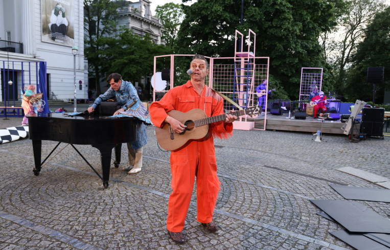 Ein Musiker in einem orangenen Overall steht auf dem Goetheplatz. Er spielt Gitarre. Hinter ihm steht ein Flügel. An dem Flügel steht ein Schauspieler. 