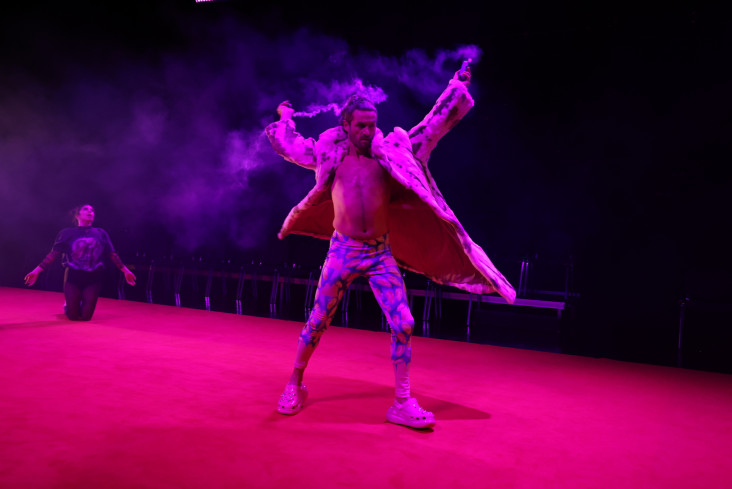 Ein Tänzer in einer bunten Leggins und einem bunten Plüschmantel dreht sich mit zwei kleinen Nebelwerfern in den Händen. Sein Mantel fliegt.