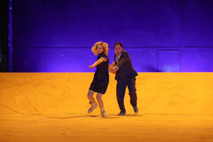 Der Bühnenboden ist gelb, der Hintergrund blau. Die Bühne ist leer. Nur eine Schauspielerin und ein Schauspieler laufen Hand in Hand auf das Publikum zu.