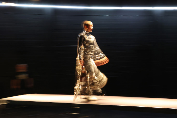 Eine Frau in einem metallisch-glänzendem Outfit geht über einen Laufsteg. Das Bild ist leicht verschwommen.