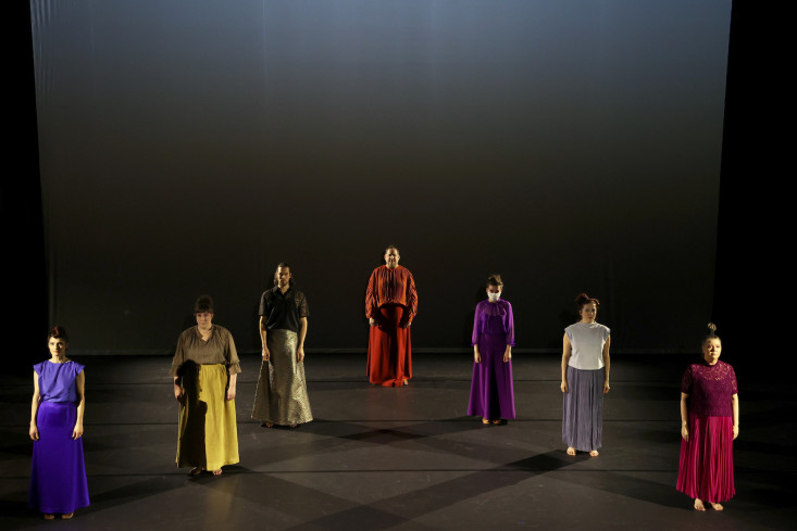 Sieben Personen stehen in dreiecksform auf der Bühne, dass die beiden äußeren ganz vorn stehen und der in der Mitte ganz hinten. Alle tragen bunte, bodenlange Röcke oder Kleider.