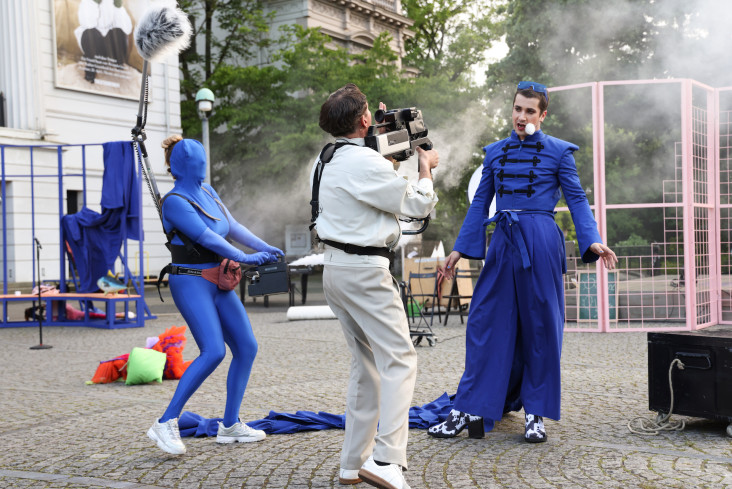 Ein Kameramann filmt einen anderen Mann. Der trägt ein auffälliges, blaues Kostüm. Neben ihnen steht eine Schauspielerin mit einer Nebelmaschine. Sie hat eine Kapuze über ihr Gesicht gezogen.