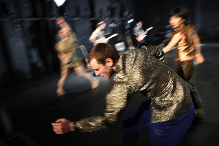 Vorn ist ein Tänzer in voller Bewegung. Er trägt eine goldene Jacke. Hinter ihm tanzen mehrere Personen. 