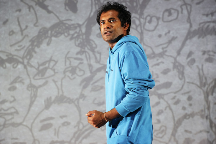 Der Schauspieler steht in einem leuchtend blauen Kapuzenpullover und schaut mit geballter Faust hoch. Hinter ihm sind auf einer weißen Leinwand viele Köpfe aufgezeichnet. Der Schauspieler steht in einem leuchtend blauen Kapuzenpullover und schaut mit geballter Faust hoch. Hinter ihm sind auf einer weißen Leinwand viele Köpfe aufgezeichnet.