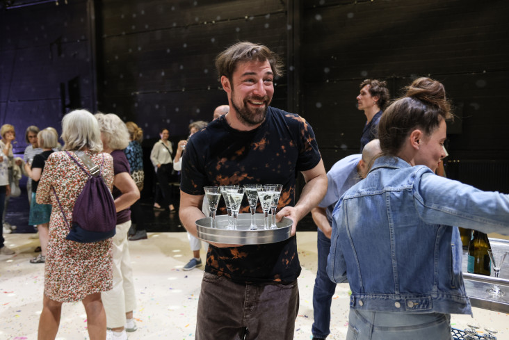 Ein Schauspieler steht mit einem Tablett auf der Bühne. Auf dem Tablett sind Sektgläser. Um ihn herum stehen viele Menschen aus dem Publikum. Alle unterhalten sich. Ein Schauspieler steht mit einem Tablett auf der Bühne. Auf dem Tablett sind Sektgläser. Um ihn herum stehen viele Menschen aus dem Publikum. Alle unterhalten sich.