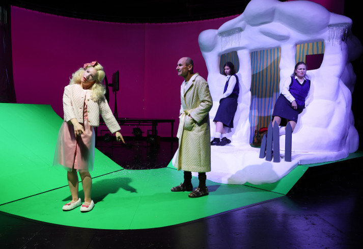 Zwei Schauspielerinnen lehnen an einem kleinen Haus. Ein Schauspieler steht weiter vorn und schaut zu einer Schauspielerin. Sie trägt eine durchsichtige Maske und eine Perücke mit Zöpfen. Zwei Schauspielerinnen lehnen an einem kleinen Haus. Ein Schauspieler steht weiter vorn und schaut zu einer Schauspielerin. Sie trägt eine durchsichtige Maske und eine Perücke mit Zöpfen.
