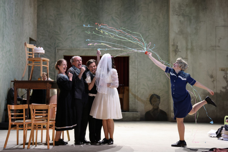 Neben ein paar aufeinander gestapelten Stühlen stehen vier Personen. Eine Schauspielerin trägt ein Hochzeitskleid und einen Schleier. Eine andere wirft Luftschlangen. Neben ein paar aufeinander gestapelten Stühlen stehen vier Personen. Eine Schauspielerin trägt ein Hochzeitskleid und einen Schleier. Eine andere wirft Luftschlangen.