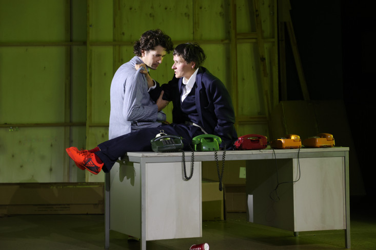 Eine Schauspielerin sitzt auf einem Schreibtisch. Sie trägt einen Anzug und neben ihr stehen fünf Telefone. Ein Schauspieler hält ihren Arm, sie umfasst seine Schulter. Eine Schauspielerin sitzt auf einem Schreibtisch. Sie trägt einen Anzug und neben ihr stehen fünf Telefone. Ein Schauspieler hält ihren Arm, sie umfasst seine Schulter.