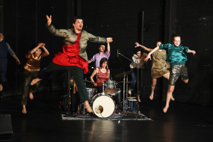 In der Mitte sitzt eine Musikerin am Schlagzeug. Um sie herum sind Tänzerinnen und Tänzer. Alle stürmen und springen nach vorn.  In der Mitte sitzt eine Musikerin am Schlagzeug. Um sie herum sind Tänzerinnen und Tänzer. Alle stürmen und springen nach vorn.