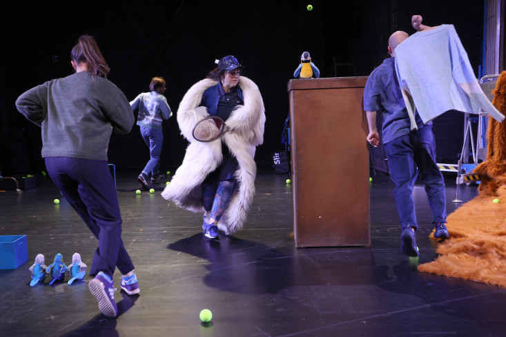 Alle Personen rennen. Drei haben dem Publikum den Rücken zugewandt. Eine Schauspielerin trägt einen Kunstfellmantel und einen Tennisschläger. Alle Personen rennen. Drei haben dem Publikum den Rücken zugewandt. Eine Schauspielerin trägt einen Kunstfellmantel und einen Tennisschläger.