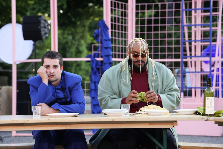 Zwei Schauspieler sitzen nebeneinander an einem Biertisch. Der eine hat einen Teller Spaghetti vor sich stehen. Er hat auch Spaghetti auf dem Kopf. Der andere stützt den Kopf in die Hand.